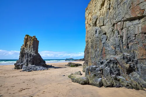 This landscape photograph captures the vibrant Godrevy rock colours on a sandy beach in Cornwall, England, during a summer afternoon. The image prominently features the striking cliffs and sea stacks that characterize this stretch of coastline in the United Kingdom. The clear blue sky and bright sunlight enhance the geological details of the weathered rock formations, while the beach leads out towards the turquoise sea. In the distance, the distinct silhouette of Godrevy Lighthouse is visible, serving as a notable landmark along the Cornish coast. The composition highlights the natural elements typical of Cornwall's beaches, focusing on the imposing cliffs and isolated sea stacks set against the scenic seaside backdrop.
